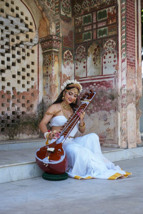 Free A woman dressed in traditional Indian attire posing with a sitar against an ancient ornamental wall. Stock Photo