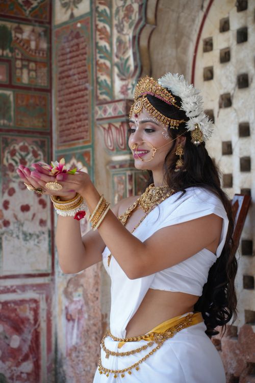 Free Indian woman dressed as a goddess in traditional attire, holding a lotus flower indoors. Stock Photo