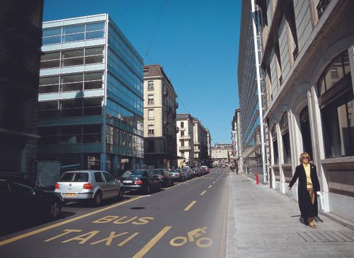 Free A city street with modern buildings, parked cars, and a pedestrian walking on a clear day. Stock Photo