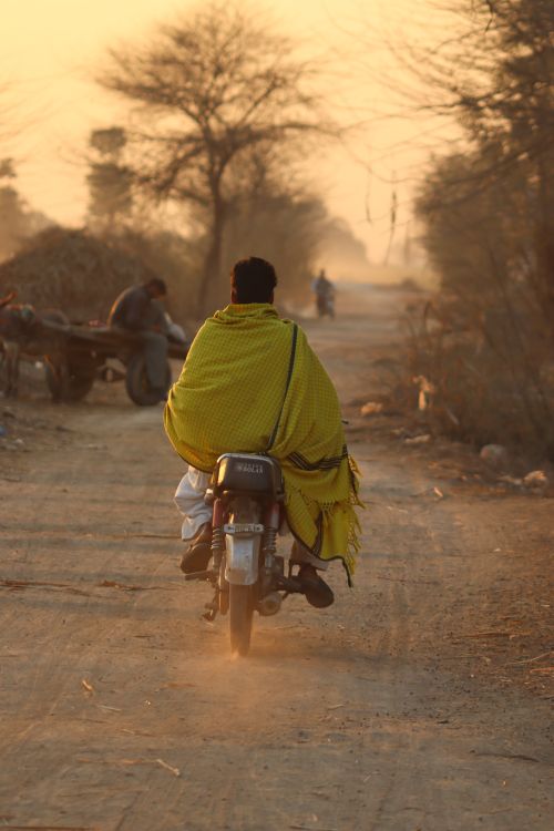 Free A person rides a motorcycle on a dusty rural path during a warm sunset, wrapped in a yellow shawl. Stock Photo