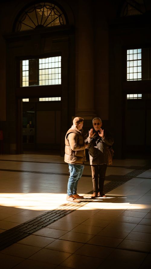 Free Two individuals conversing in a sunlit indoor atrium with striking shadows. Stock Photo