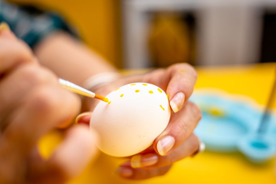Creative close-up of a person painting a white Easter egg with yellow dots, capturing the art and craft of Easter decoration.