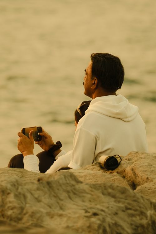 Free A man in a white hoodie sits on rocks, enjoying a tranquil sea view at sunset. Stock Photo
