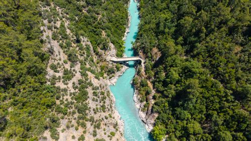 Free Aerial perspective of a turquoise river winding through lush green forested hills with a stone bridge crossing. Stock Photo
