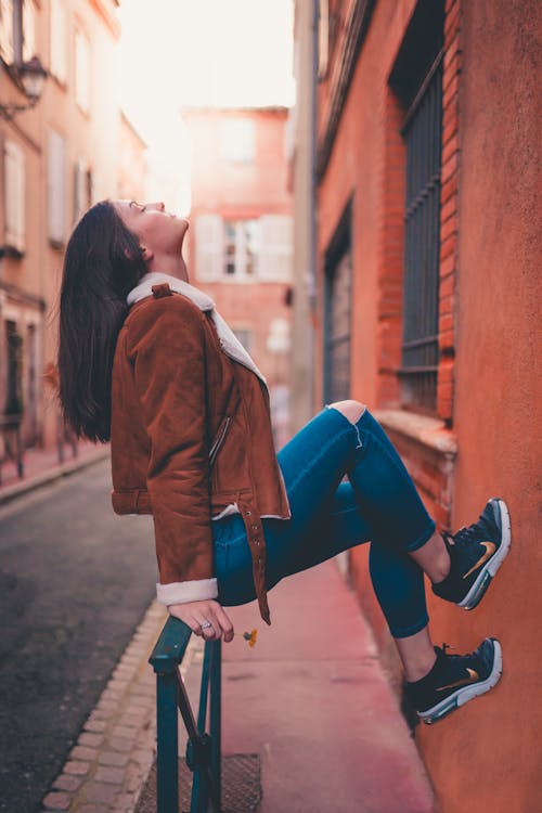 Free Woman In Brown Jacket And Blue Denim Jeans Sitting On Handrail Stock Photo
