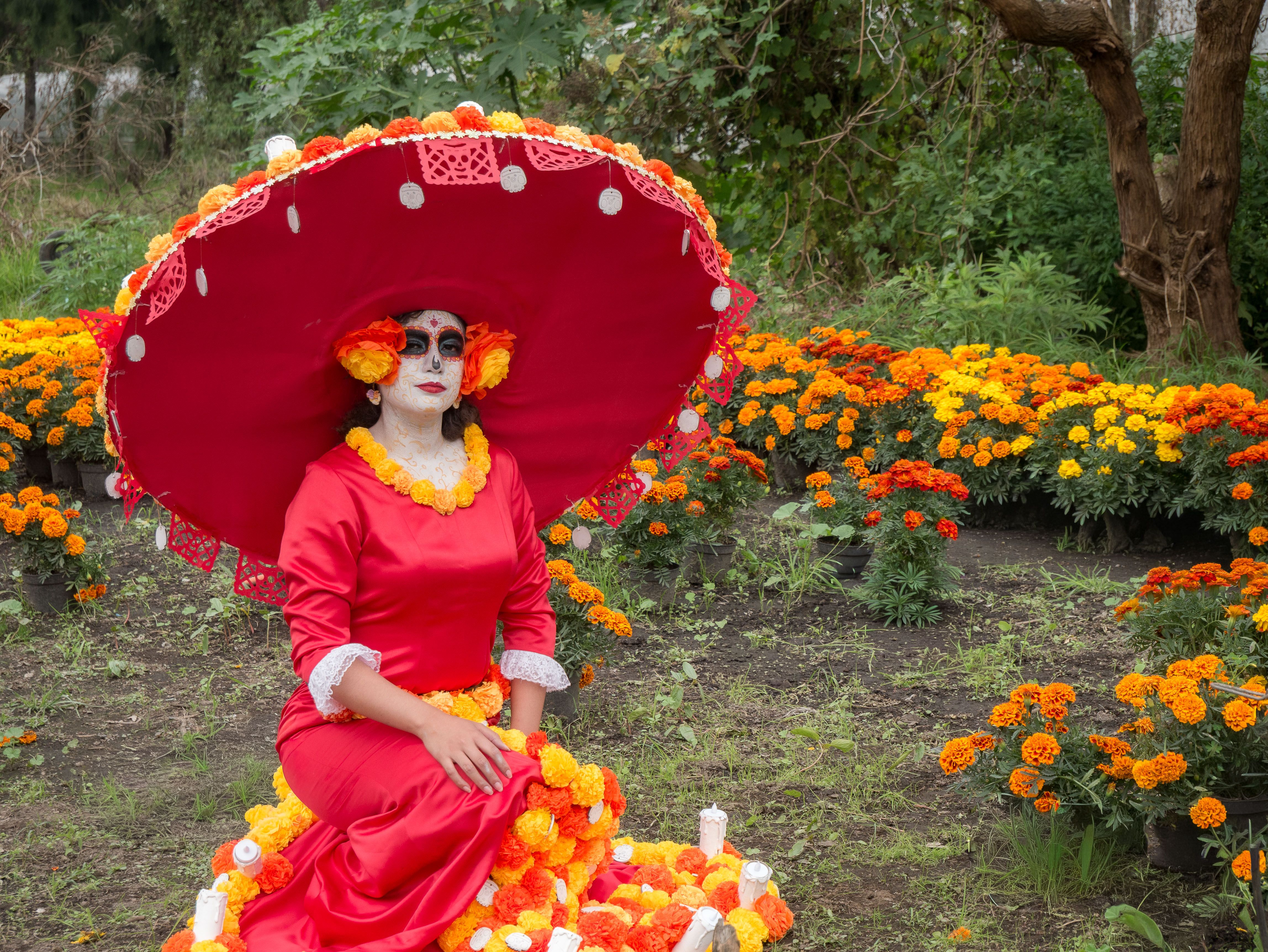 A woman dressed as La Catrina, adorned in vibrant colors, sits among marigolds for Dia de los Muertos.