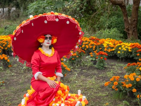 A woman dressed as La Catrina, adorned in vibrant colors, sits among marigolds for Dia de los Muertos.