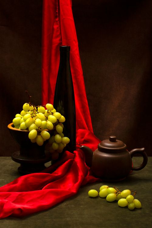Free A sophisticated still life composition featuring green grapes, a black bottle, and a teapot against a red drape. Stock Photo