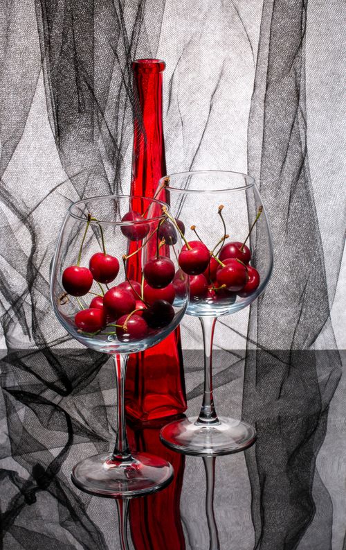 Free Classic still life featuring red cherries in wine glasses with a red vase on a reflective surface. Stock Photo
