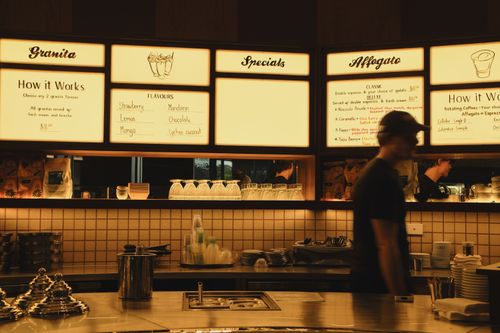 Free A warm, inviting cafe scene with glowing menu signs and a silhouetted barista in motion. Stock Photo