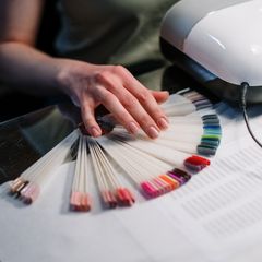 Hand with pale pink nails choosing from a fan of gel colour swatches beside a white nail lamp