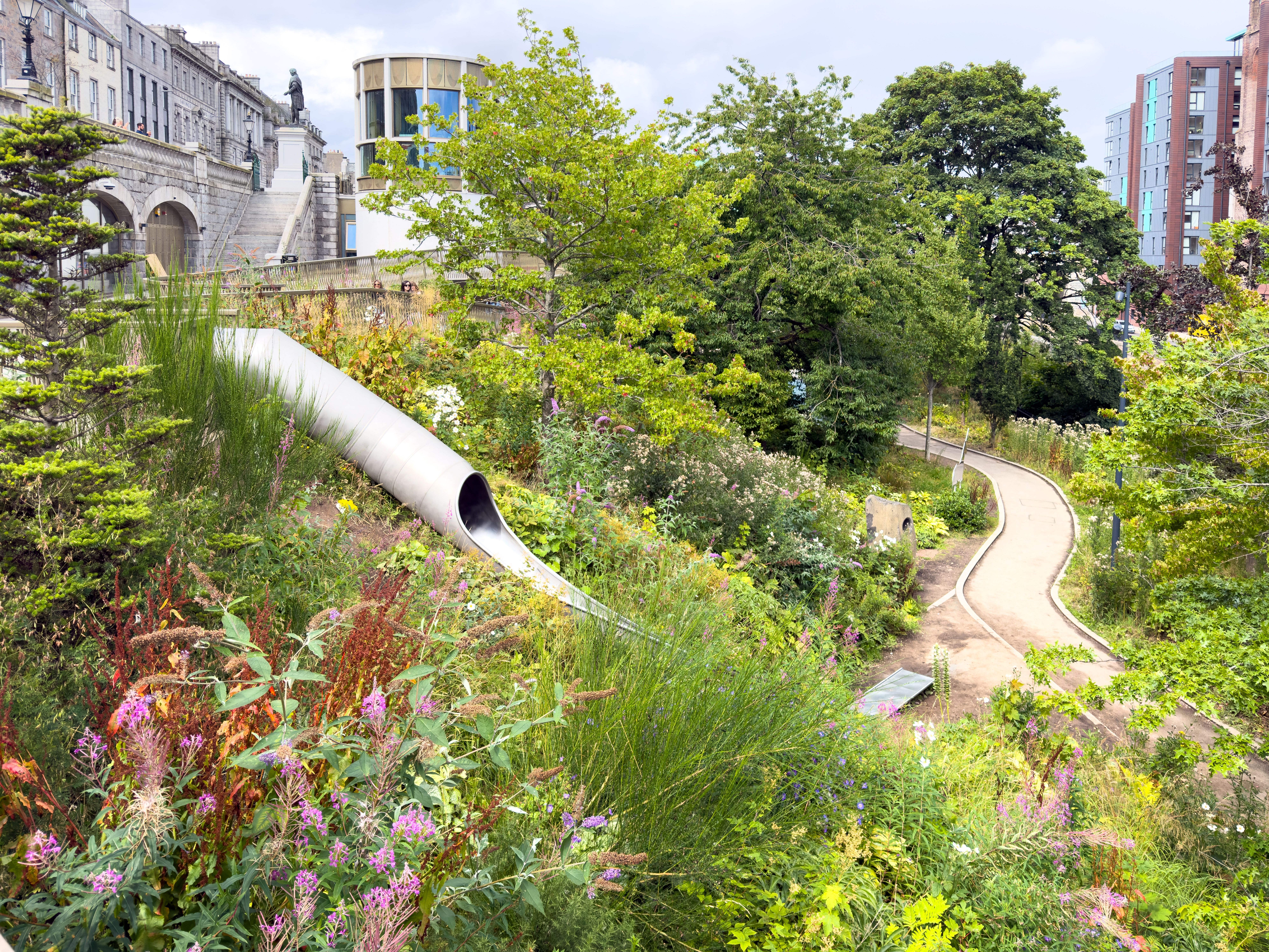 Lush greenery surrounds a slide and curved pathway in an urban park setting, ideal for relaxation.