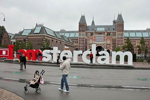 Tourists near the I Amsterdam sign with Rijksmuseum in the background on a cloudy day.