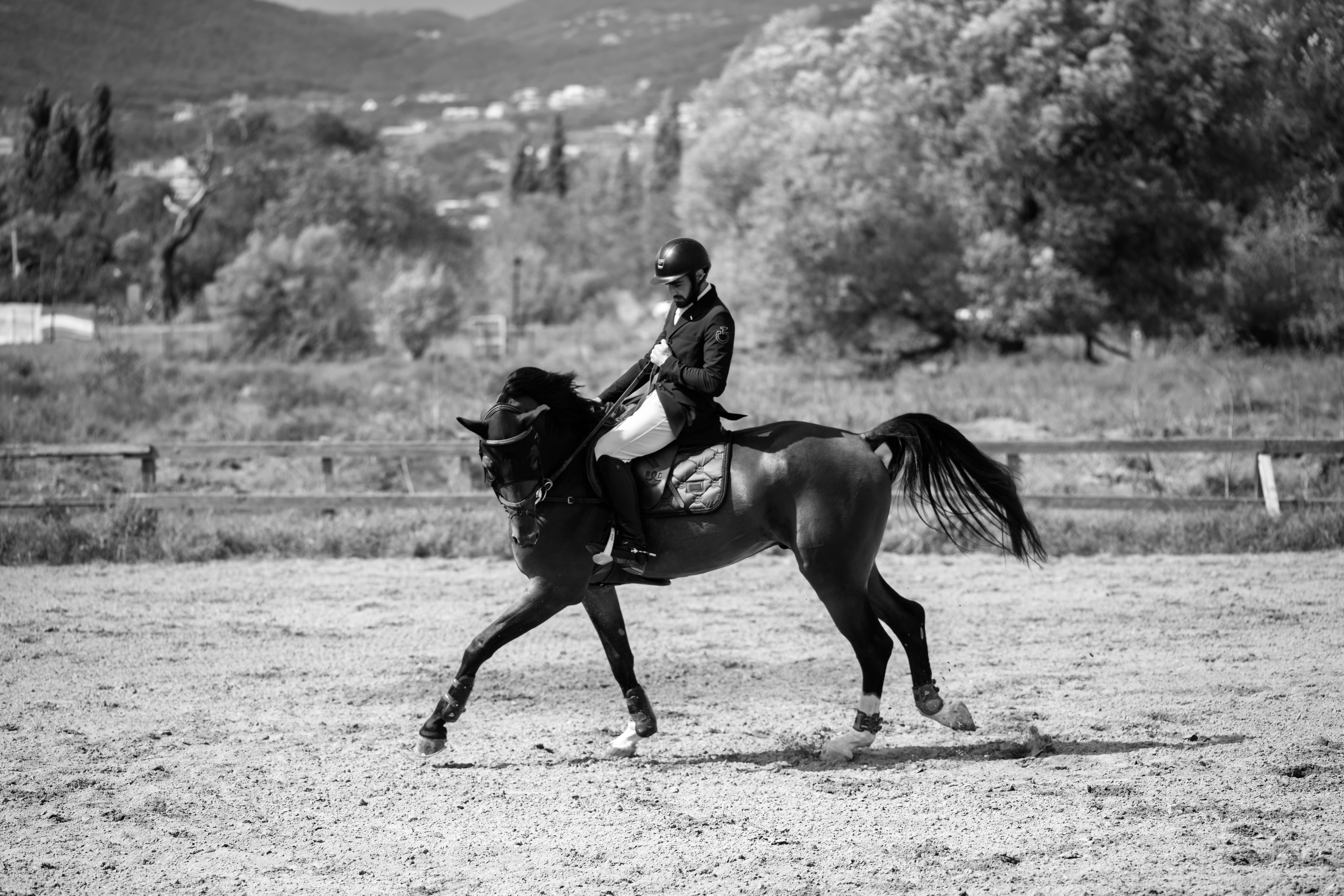 Black and white photo of an adult show jumper riding a horse in an outdoor arena with scenic mountain backdrop.