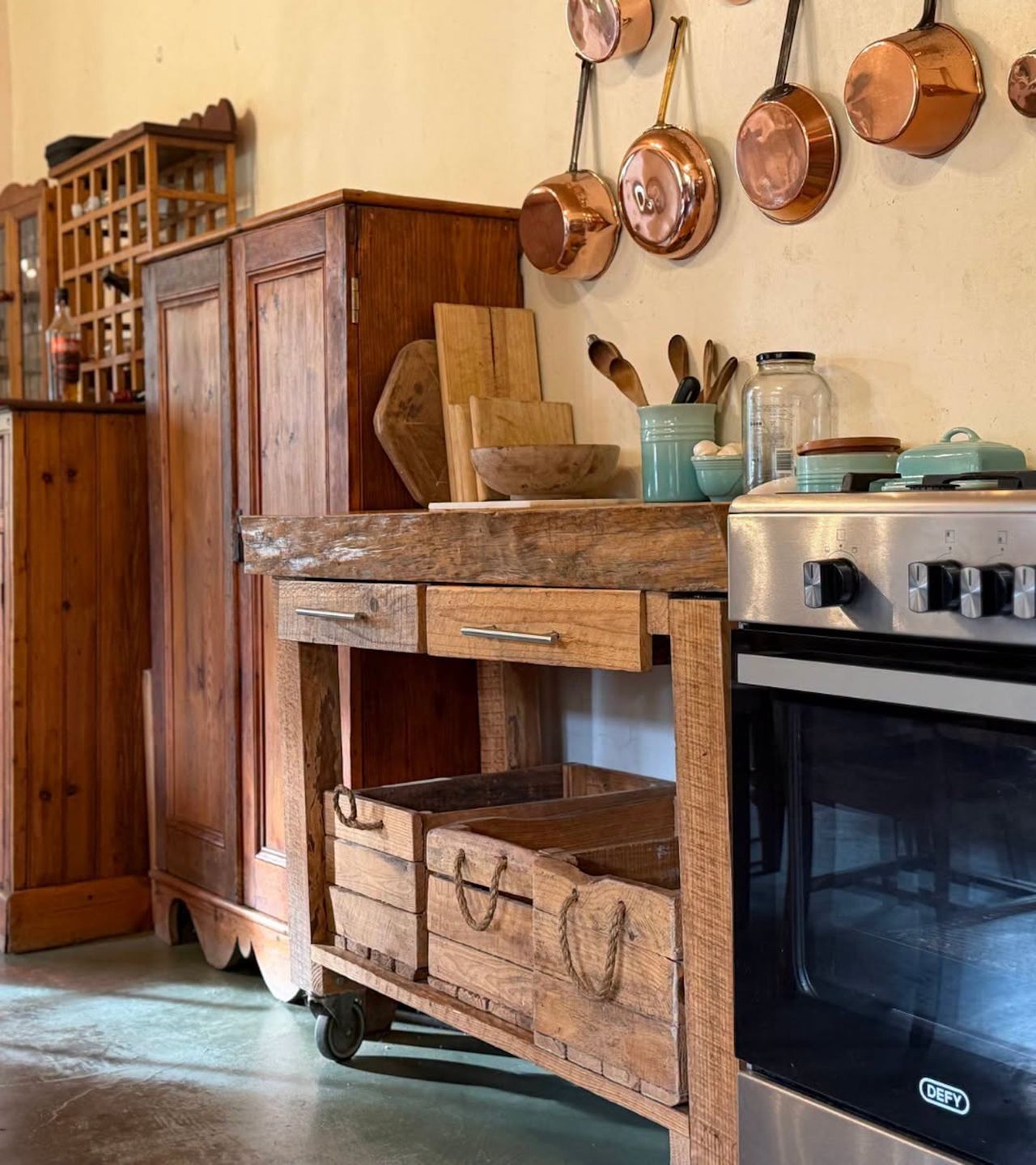 Rustic wood worktable with storage crates beside a stove, copper pots hanging above