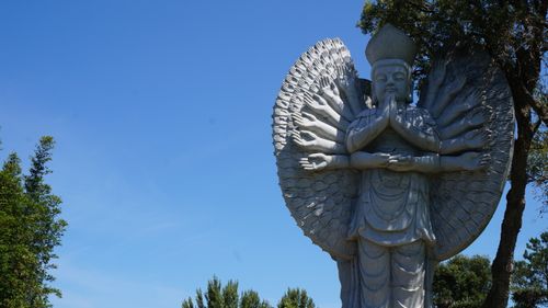 Free Captivating stone statue with multiple arms under a clear blue sky, surrounded by lush greenery. Stock Photo