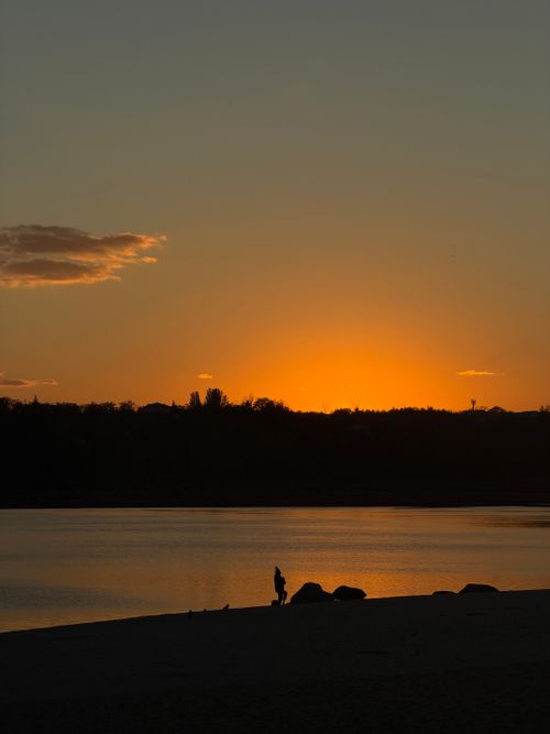 Free Silhouetted person standing by a tranquil lake during a vibrant sunset, creating a serene atmosphere. Stock Photo