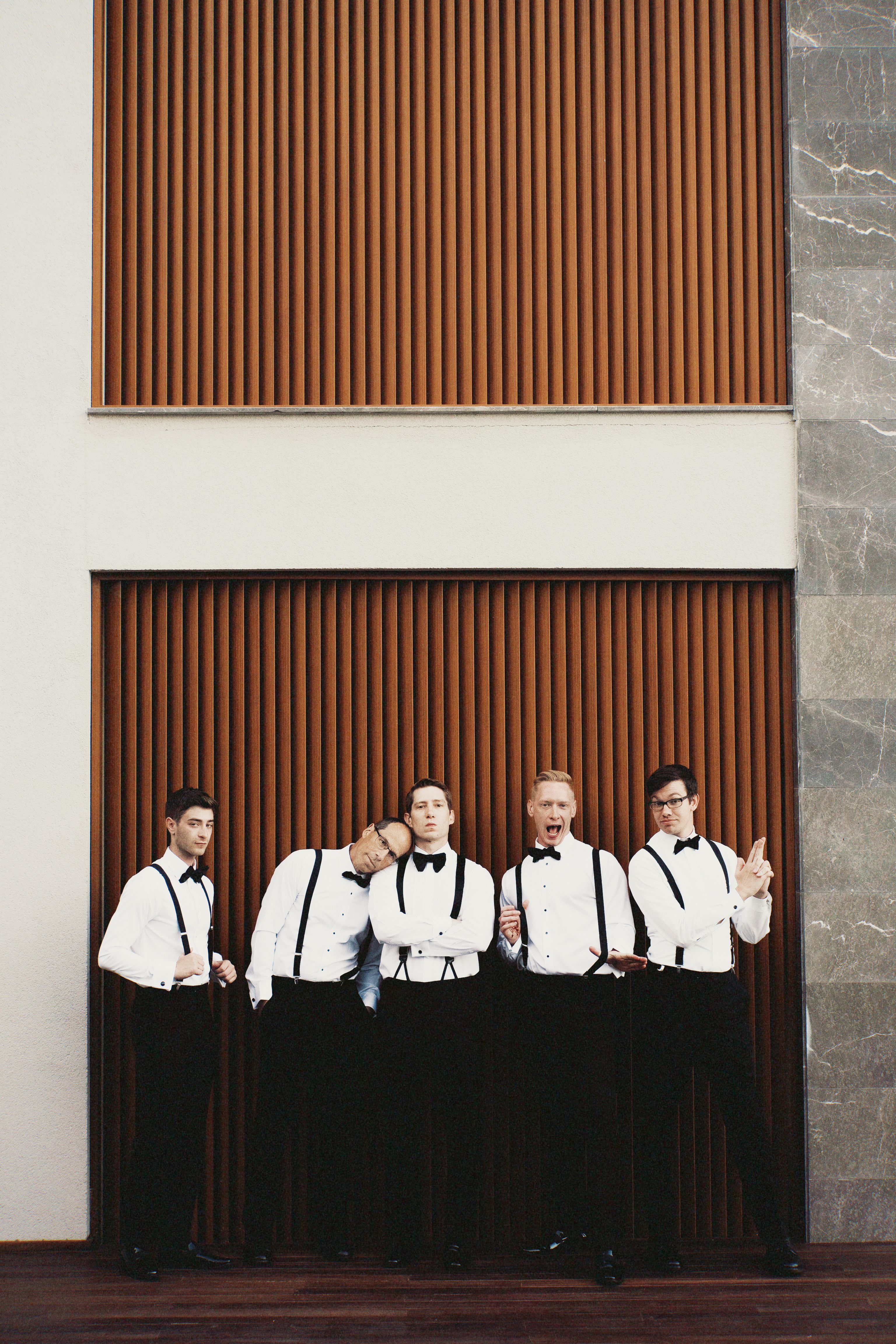 Five men in formal attire posing playfully against a modern wall.