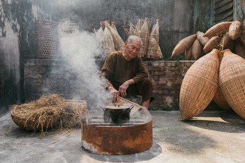 Free An elderly man in Vietnam is crafting traditional fish traps amidst smoke outdoors, showcasing cultural heritage. Stock Photo