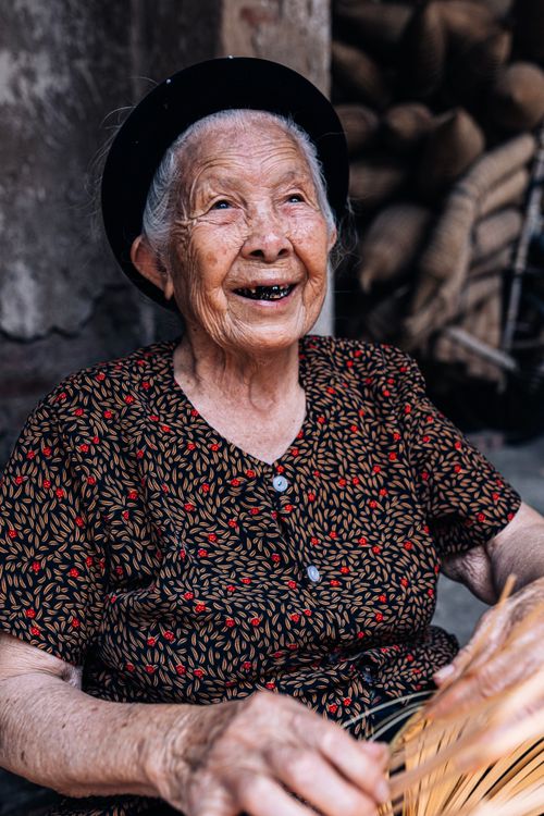 Free Candid portrait of a joyful senior Vietnamese woman weaving a basket outside. Stock Photo
