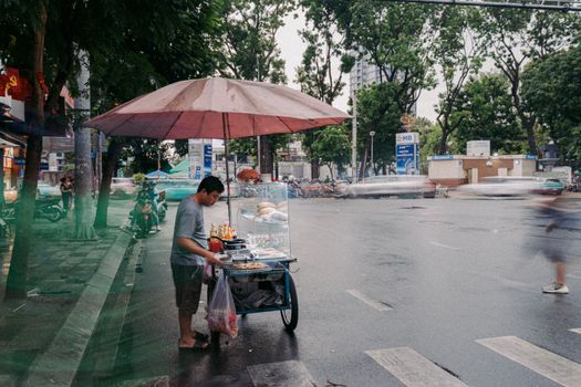 A Vietnamese street vendor selling food under an umbrella on a rainy city street.