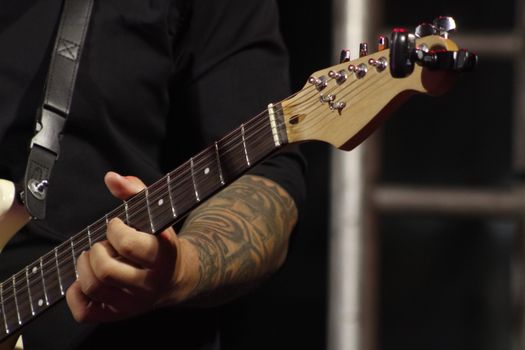 Musician's hand strumming an electric guitar on stage, showcasing tattoo and guitar details.