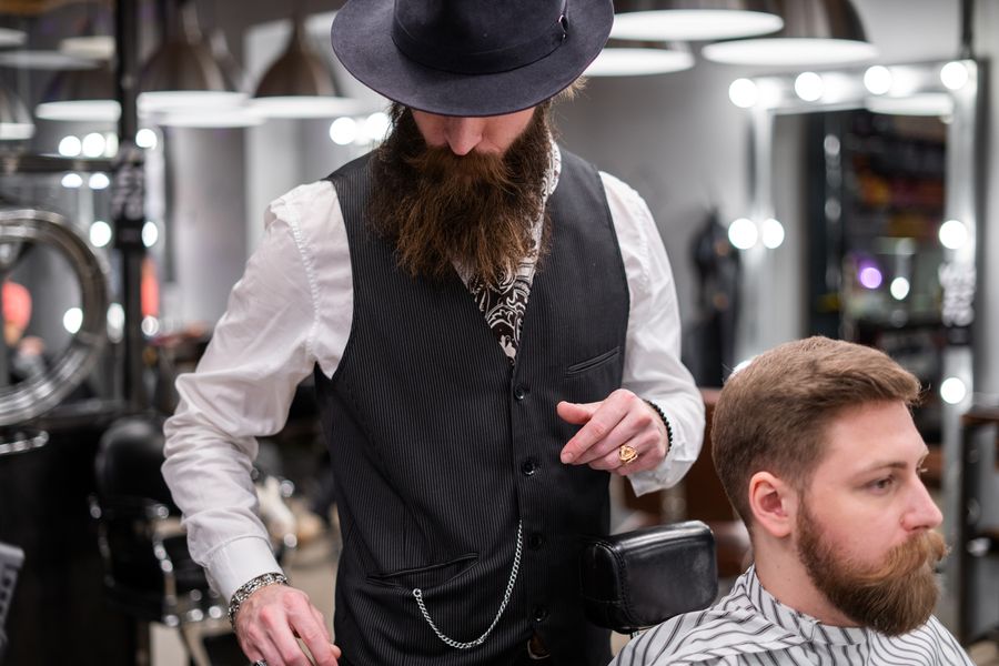 A barber in a waistcoat holding clippers beside a seated customer