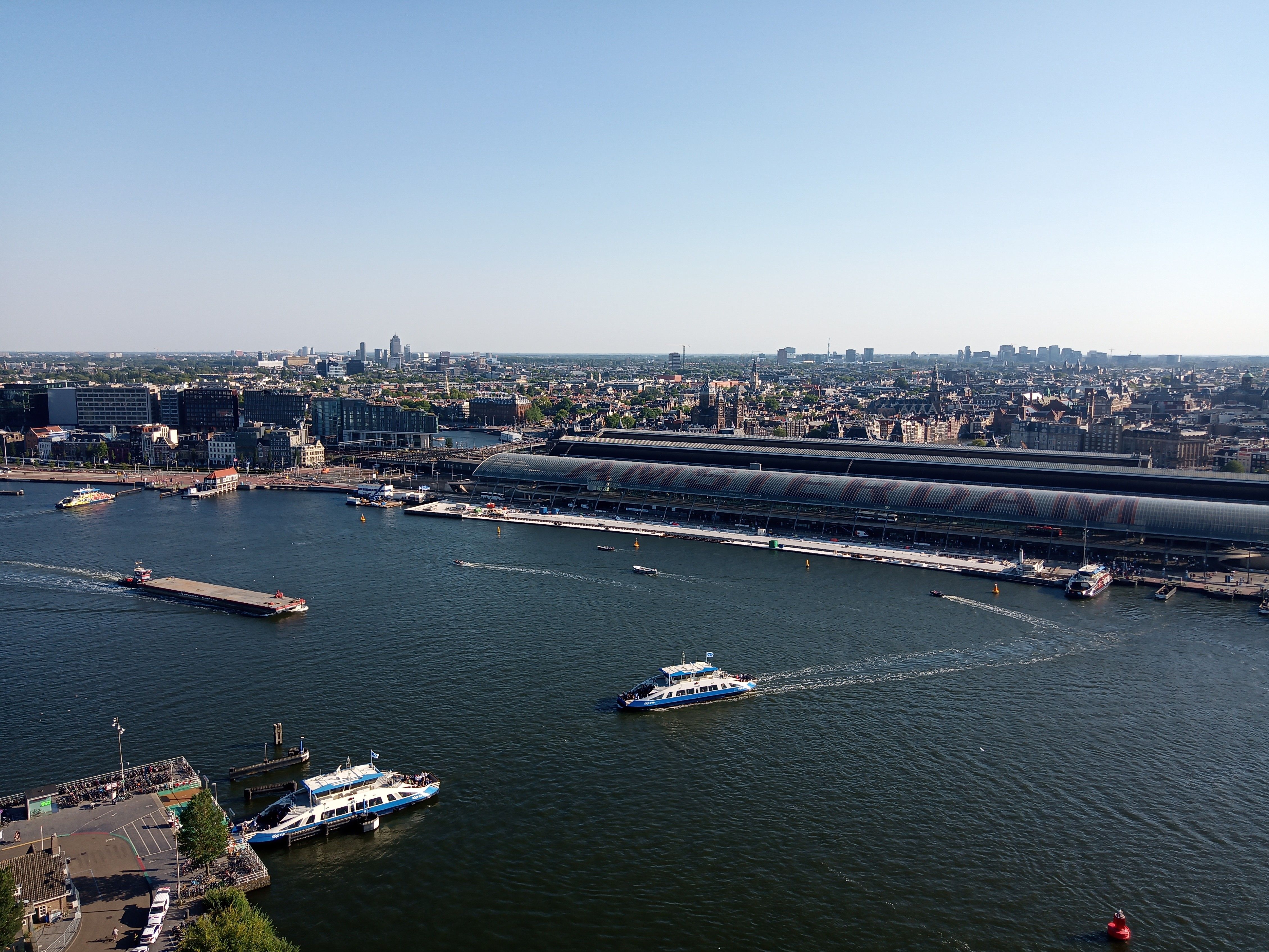 Aerial view of Amsterdam Central Station beside the canal on a sunny day, showcasing the city's architecture and vibrant waterways.