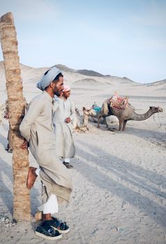 Men in traditional attire rest by camels in a sandy desert landscape under a bright sky.