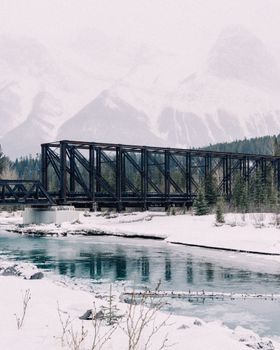 Tranquil winter landscape capturing Canmore's snow-covered bridge and river with mountainous backdrop.