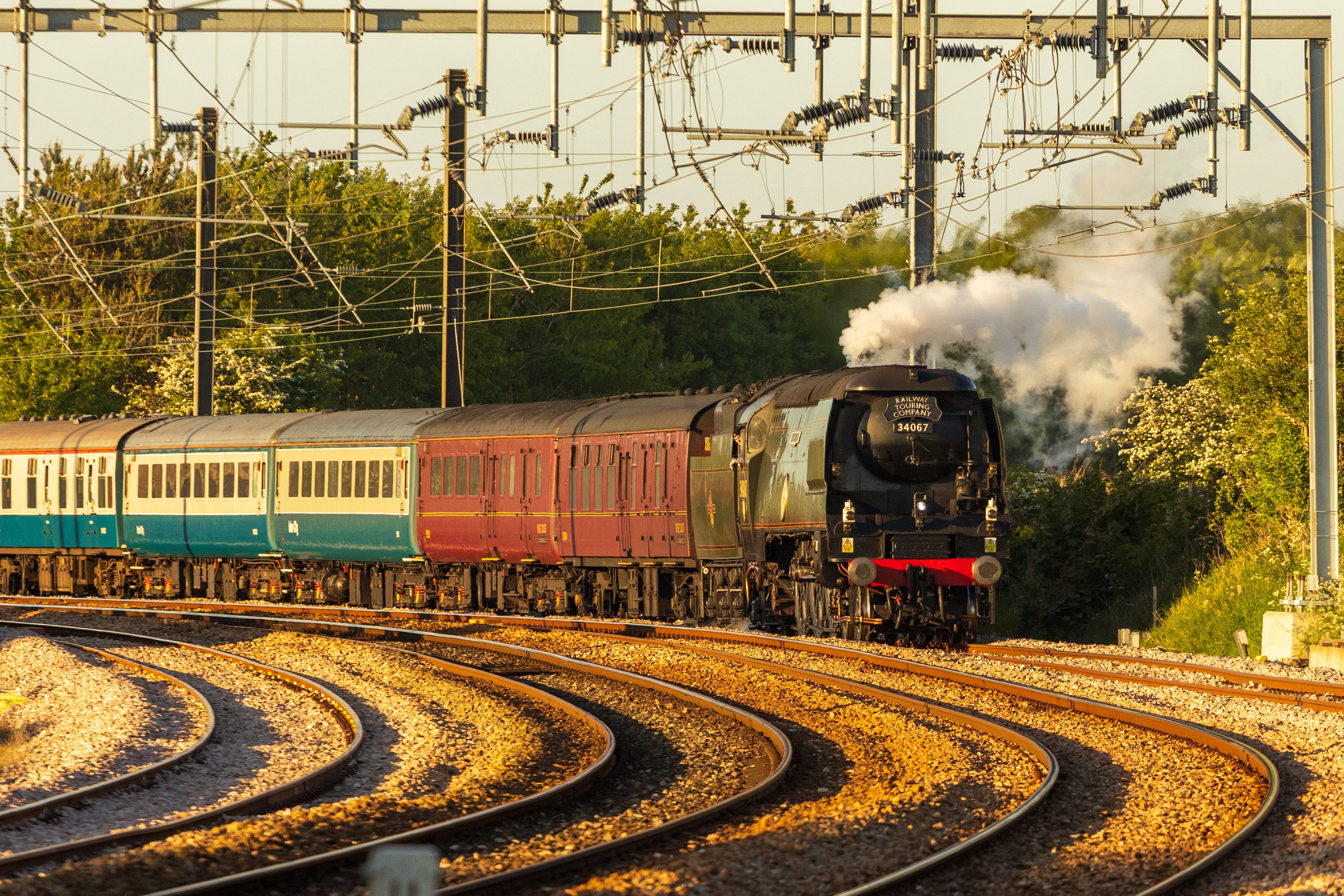 A vintage steam train travels on a curving railway track on a sunny day surrounded by greenery.