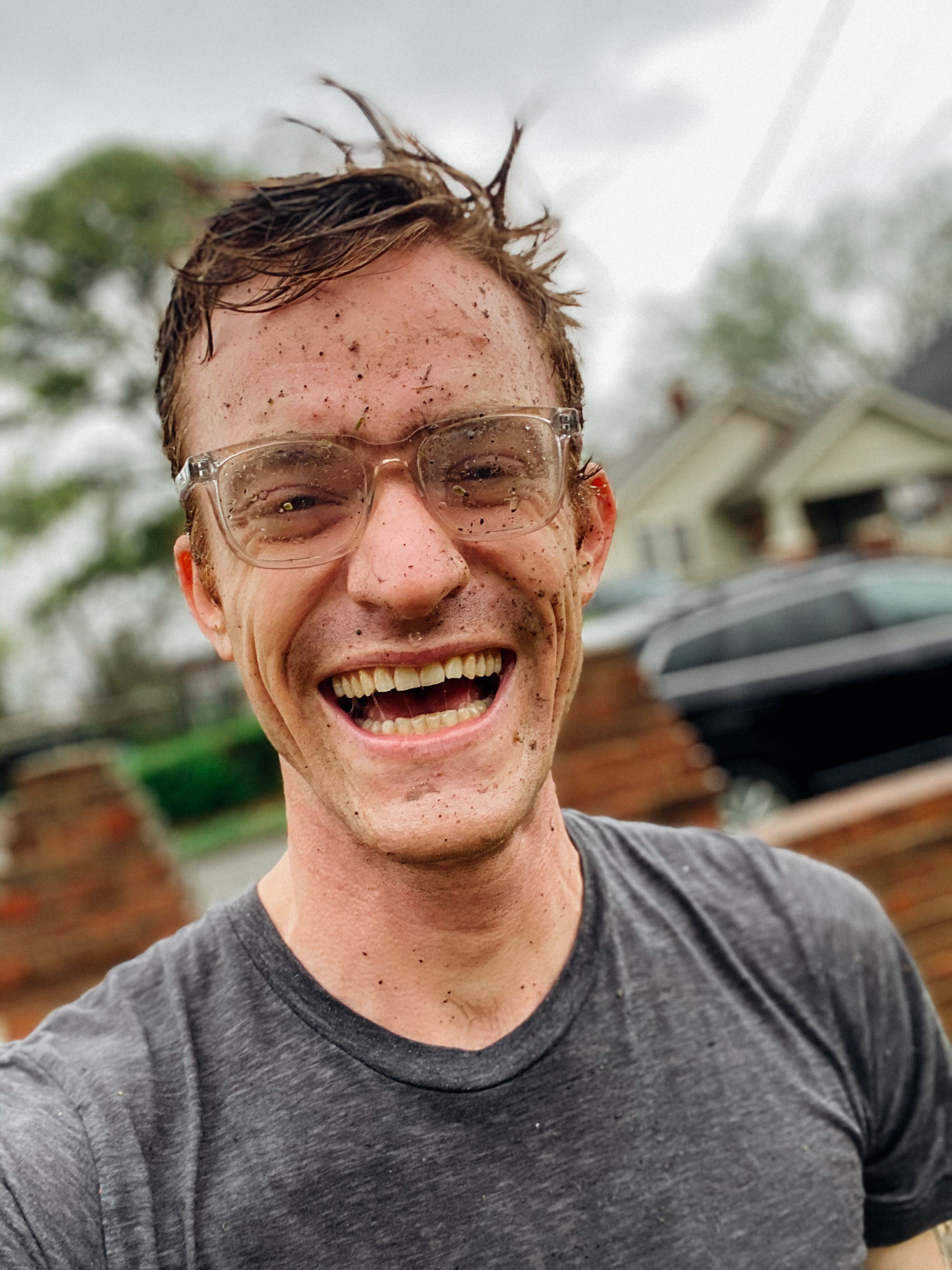 Smiling young man with muddy face and glasses standing outdoors in a casual t-shirt.