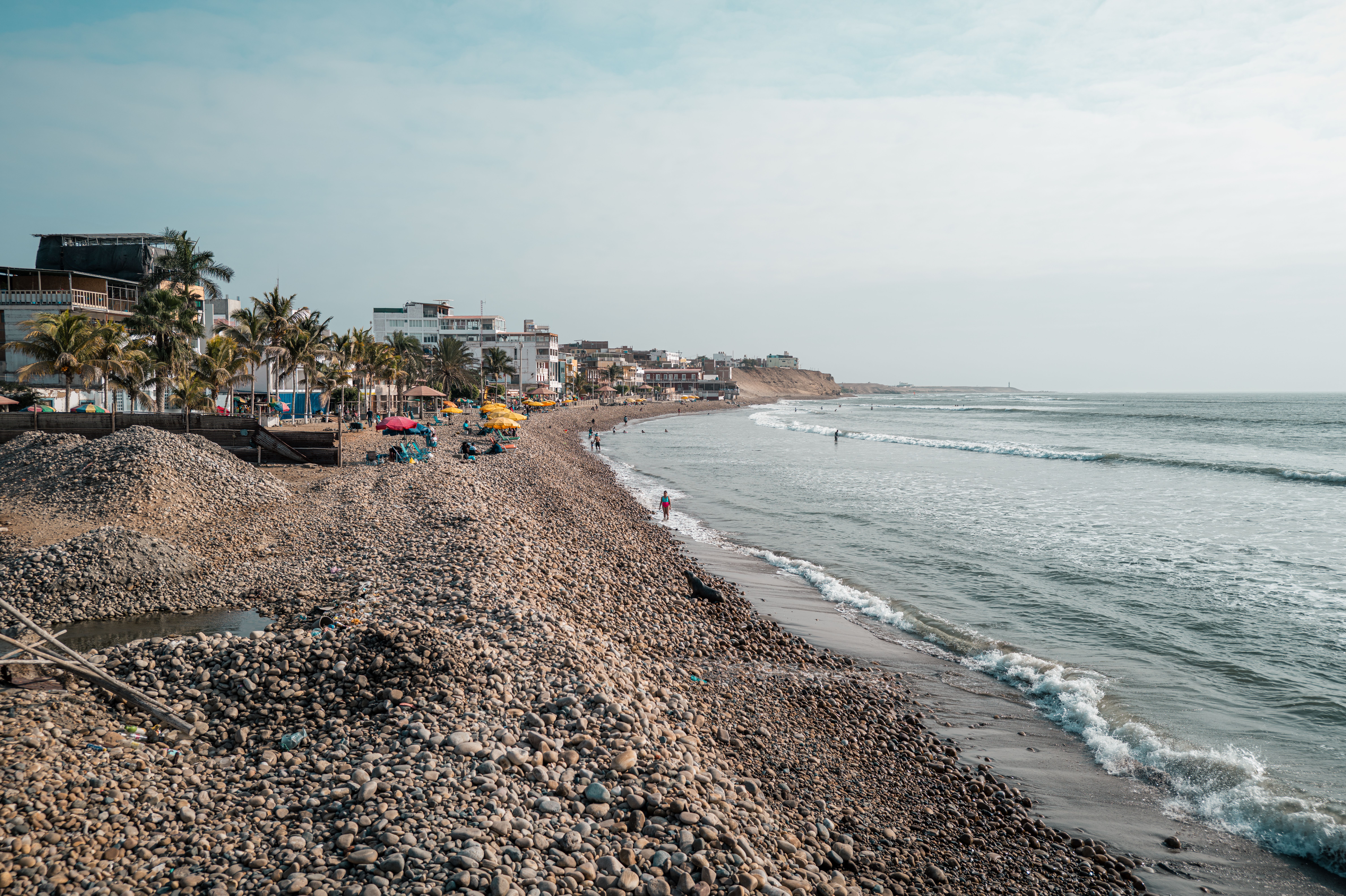 Pebble beach with waves and buildings in Pacasmayo, Peru, under a clear sky.