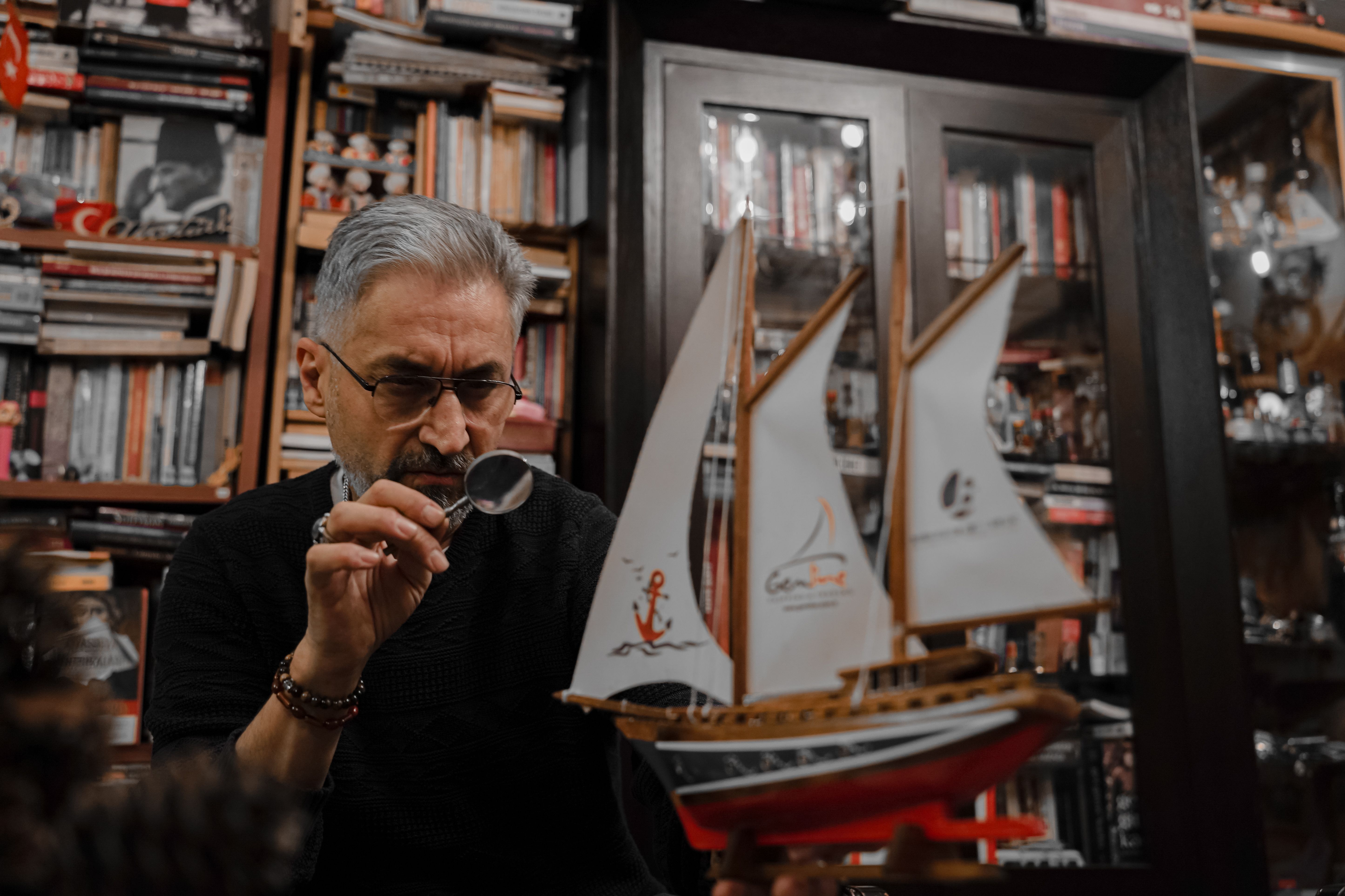 Temsilî fotoğraf: Man examines a model ship using a magnifying glass in a book-filled room, displaying focus and hobby enthusiasm.