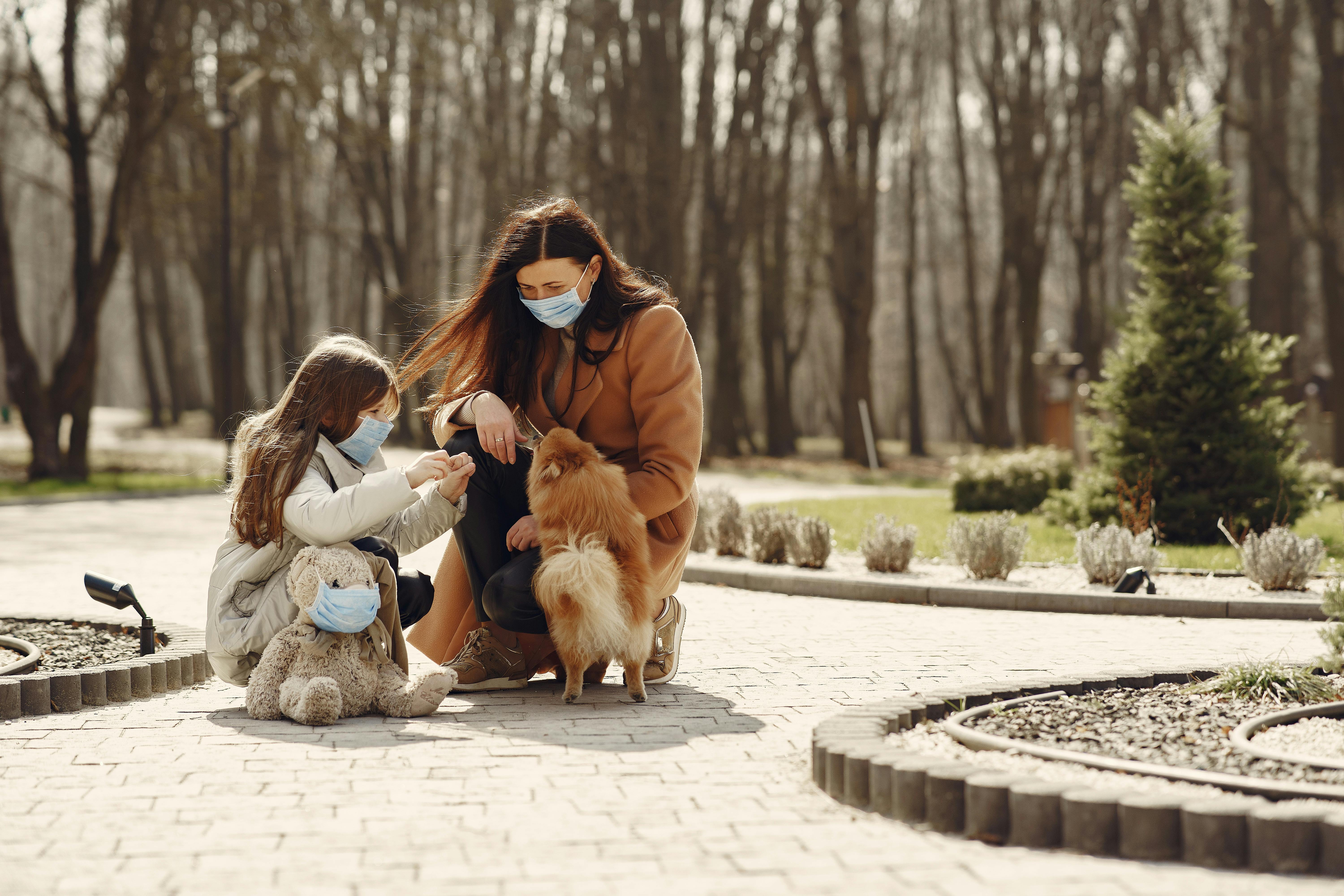 happy woman with cute girl wearing protective mask and playing with adorable dog in park alley