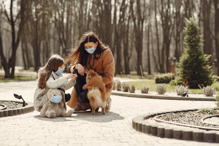 Happy Woman With Cute Girl Wearing Protective Mask And Playing With Adorable Dog In Park Alley