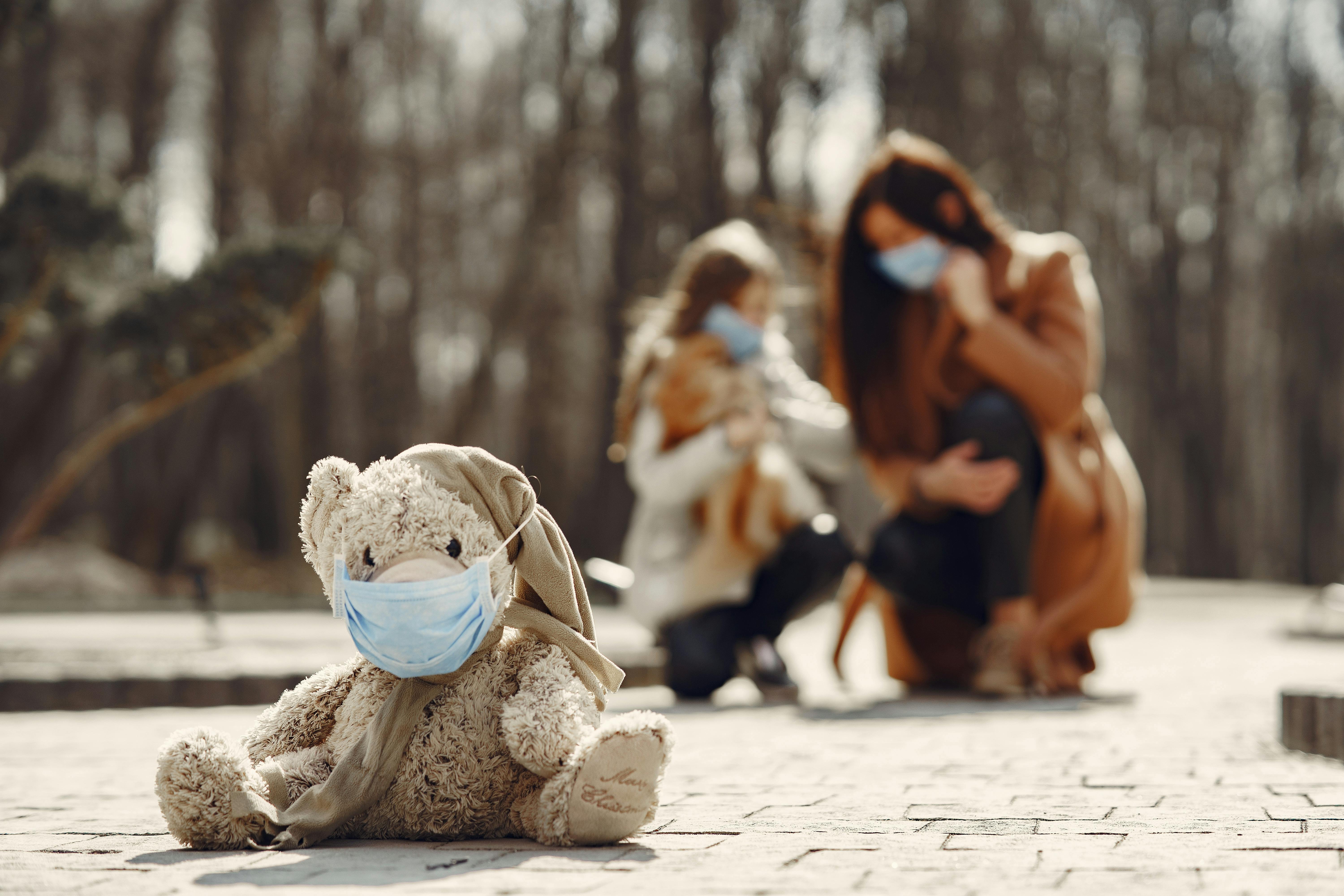 shabby teddy bear in medical mask sitting on pavement against blurred mom and kid in park