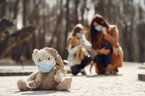 Cute children toy in scarf and mask placed on asphalt against blurred owners woman and girl in warm wear playing in nature