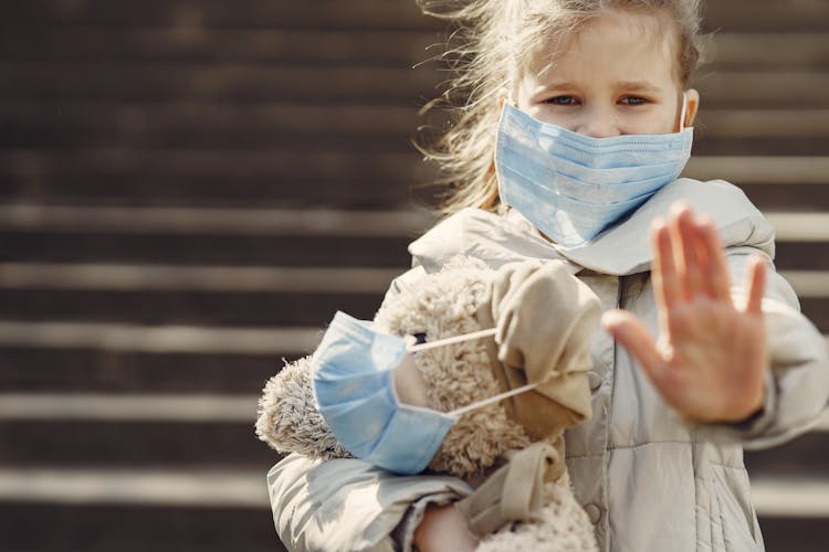 Serious Girl In Protective Mask Holding Plush Toy In Mask And Showing Palm Against Steps