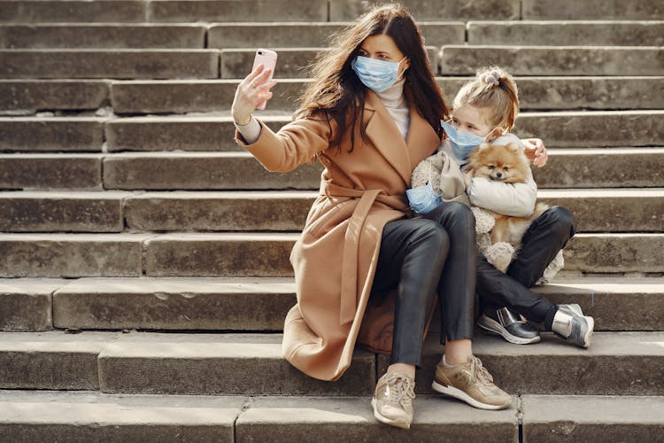 Young Woman Hugging Cute Daughter With Dog In Masks Taking Selfie On Steps