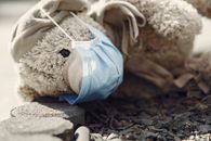 Cute plush toy in scarf and medical mask placed on ground near small sharp pebbles in street at daytime