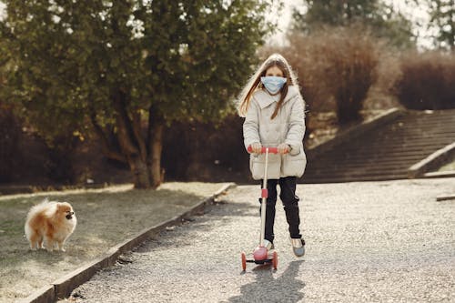 Full length of little girl in mask riding scooter while walking pet in nature