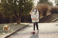 Full length of little girl in mask riding scooter while walking pet in nature