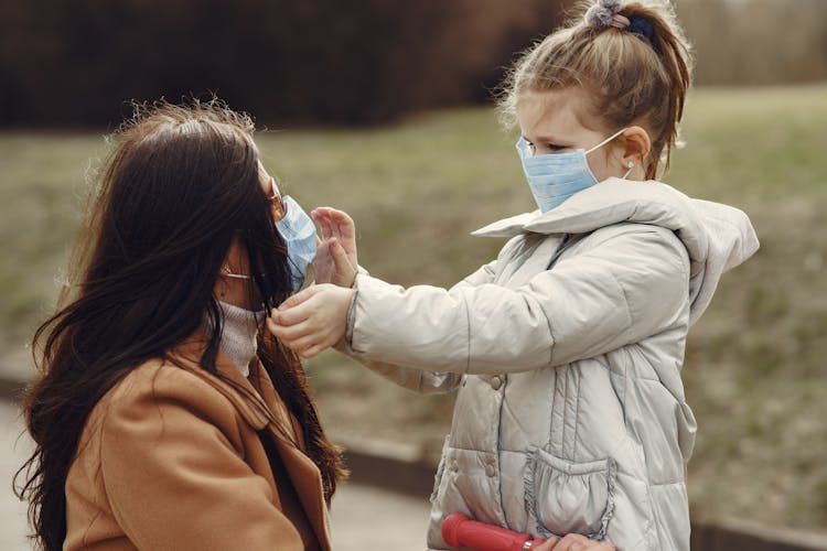 Cute Little Girl In Mask Helping Put On Medical Mask For Mom In Sunglasses During Stroll In Park