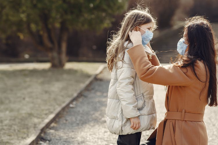 Young Woman And Child Helping Each Other Ti Wear Disposable Masks In Nature