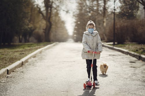 Full body full length of cute little girl in disposable mask riding scooter near funny dog running on pavement in autumn forest