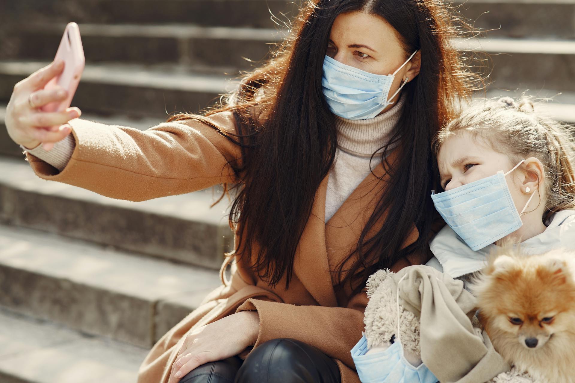 Young woman and cute girl with toy wearing masks and taking selfie while chilling with dog on steps