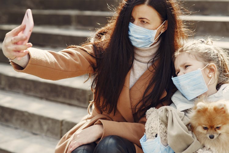 Young Woman And Cute Girl With Toy Wearing Masks And Taking Selfie While Chilling With Dog On Steps