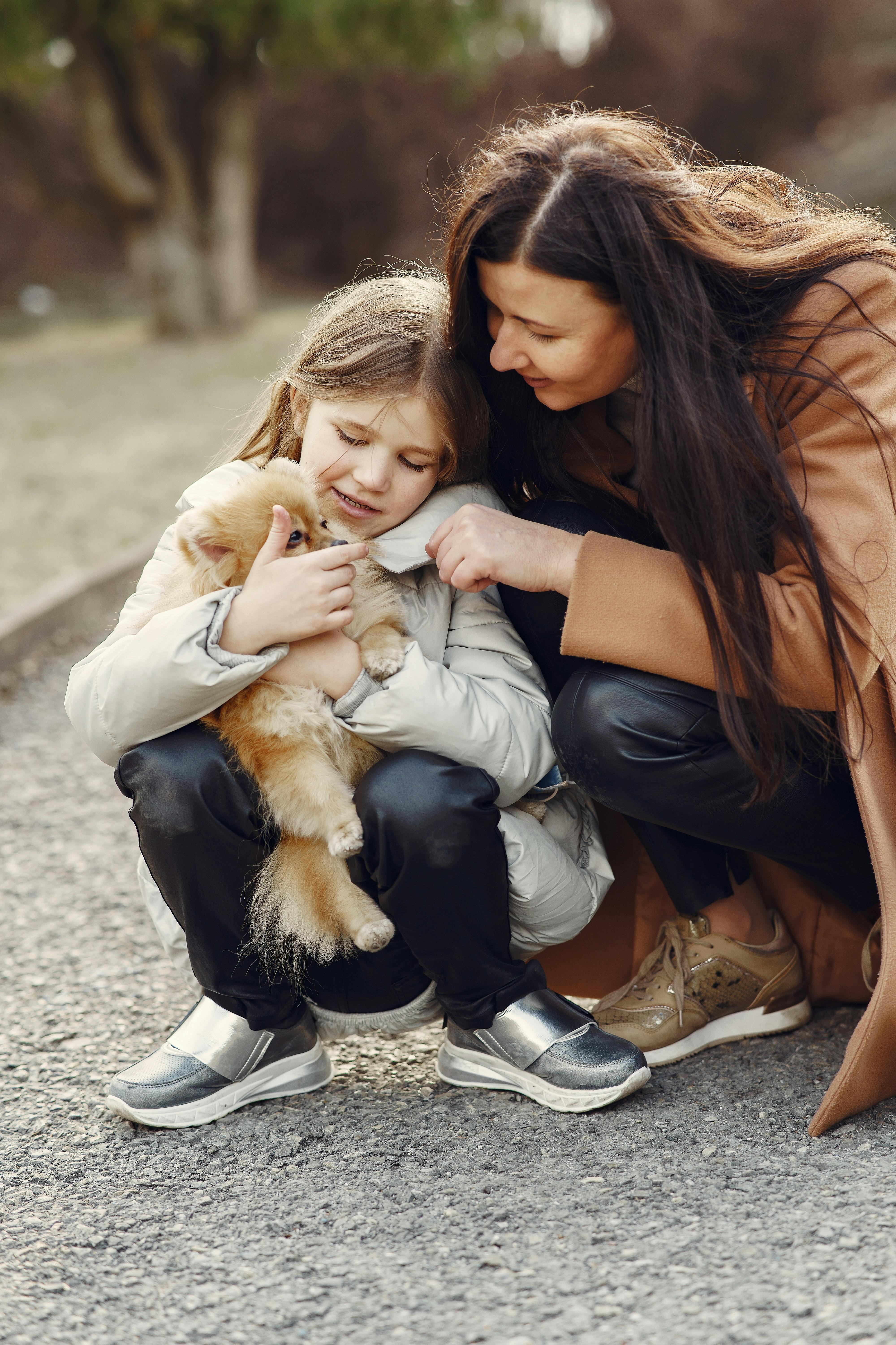 From above of cheerful young woman and little girl wearing warm wear squatting together and embracing fluffy Pomeranian dog while resting in nature
