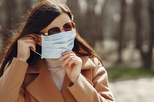 Casual female in warm coat and sunglasses putting on medical mask during stroll in autumn park at daytime against blurred background